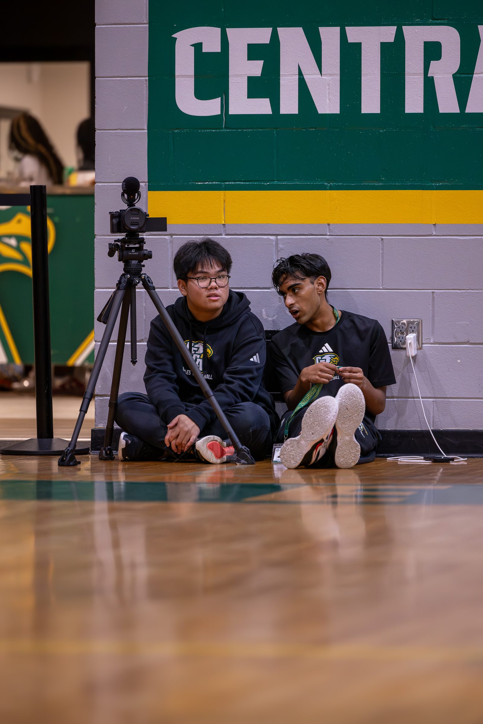Two student crew members seated on the floor beside a camera on a tripod at a live athletics event